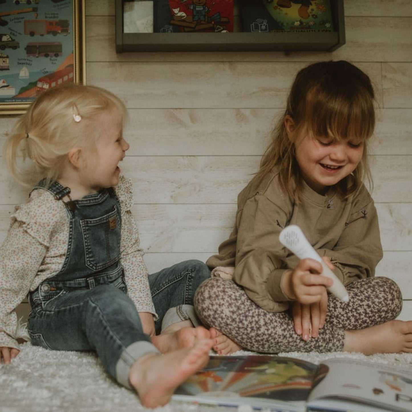 Två barn sitter på golvet och skrattar medan de använder Readioo läspenna med boken Bokstäver.