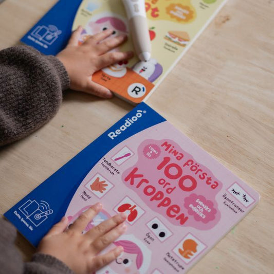 Barnhand som pekar på interaktiv bok "Mina första 100 ord: Kroppen" från Readioo tillsammans med Readioo läspenna.