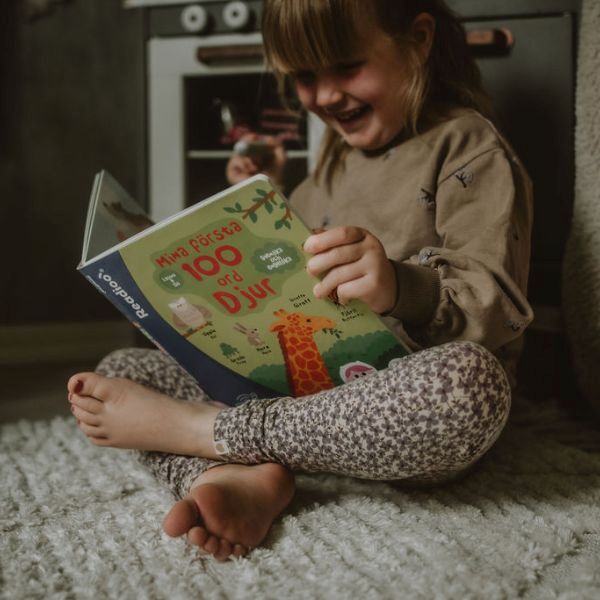 Flicka sitter på mattan och läser Readioo-boken "Mina första 100 ord: Djur".