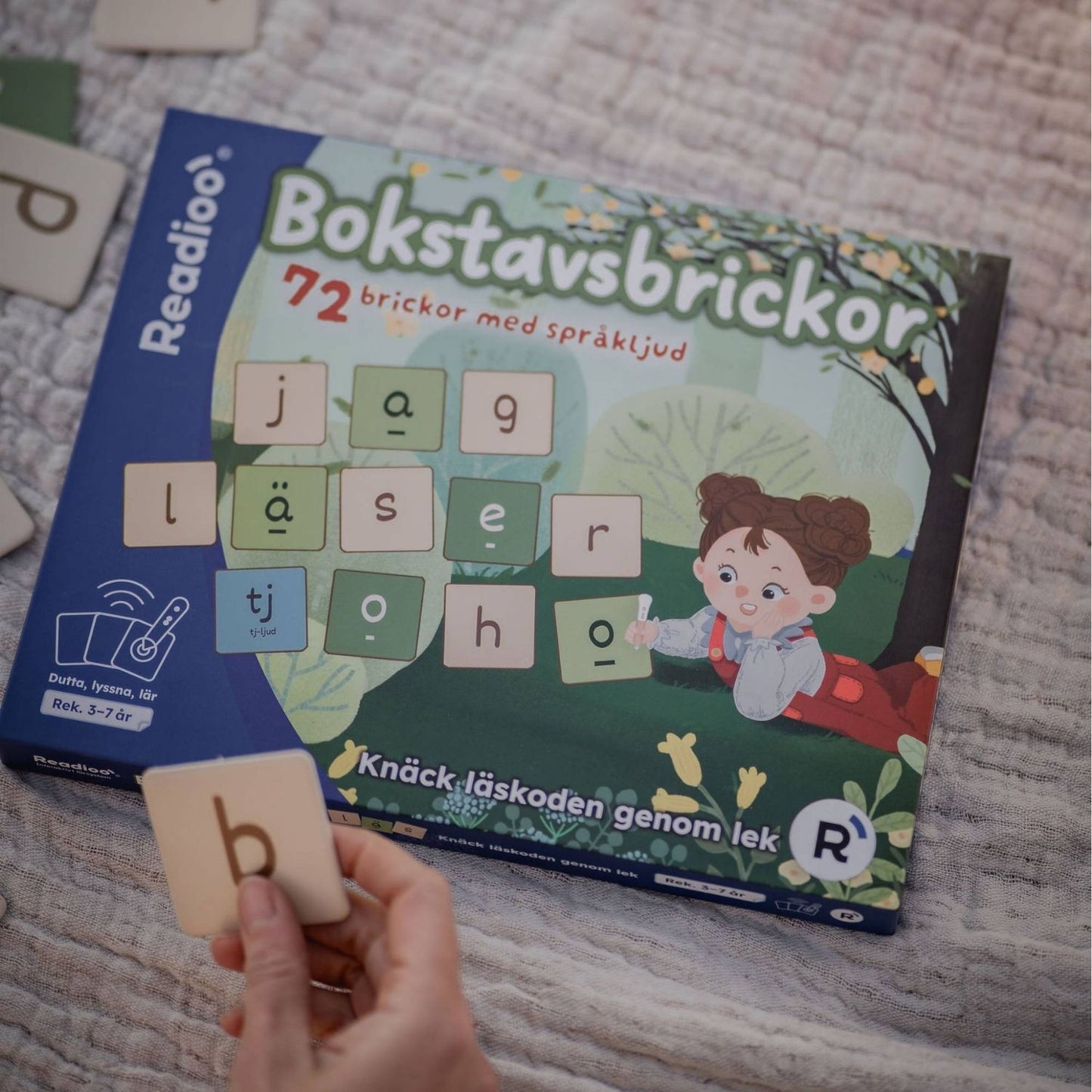 Readioo Bokstavsbrickor, förpackningen på ett tyg med en hand som håller upp en bokstavsbricka med bokstaven b.