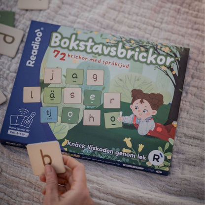 Readioo Bokstavsbrickor, förpackningen på ett tyg med en hand som håller upp en bokstavsbricka med bokstaven b.