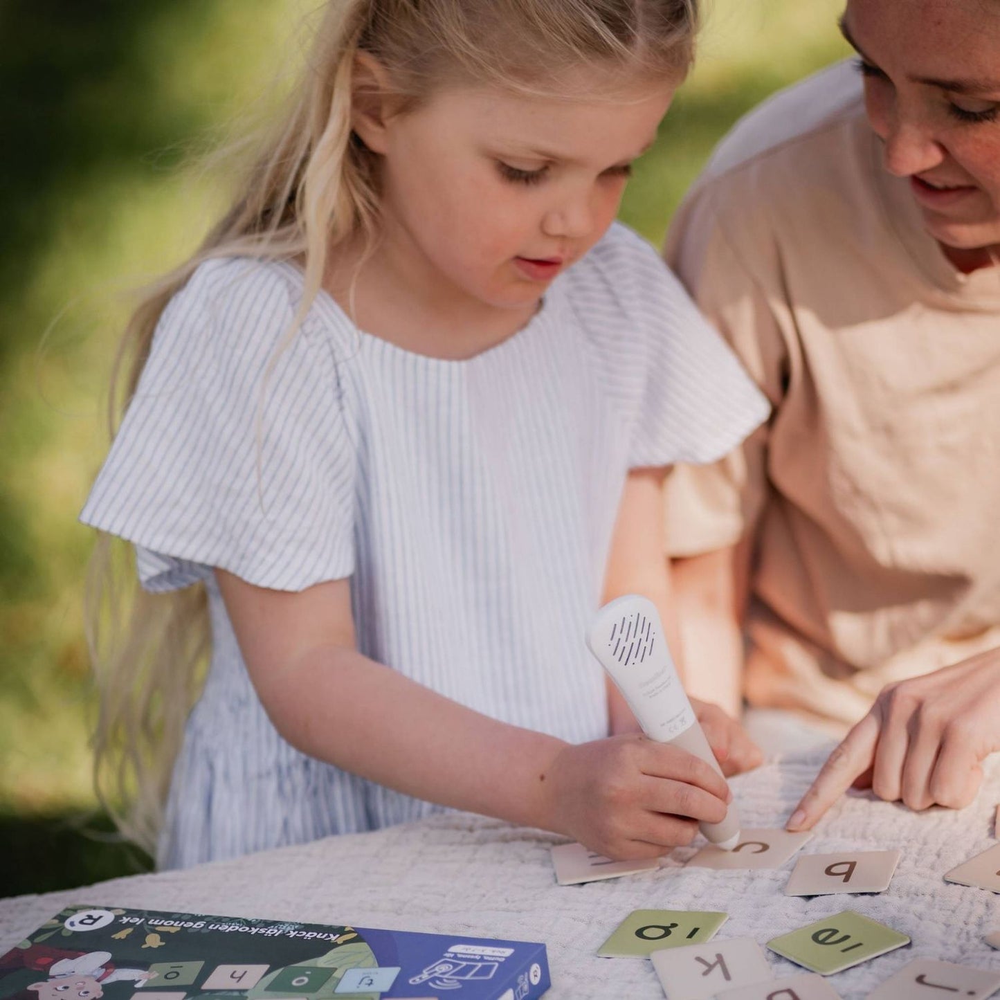 Barn använder Readioos läspenna på bokstavsbrickor utomhus, med en vuxen bredvid som pekar på en av brickorna.