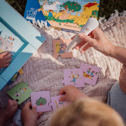 Barn och vuxen pusslar ihop bitar från Pippi Långstrump Pusseltåg på en filt utomhus, med läspennan i handen.